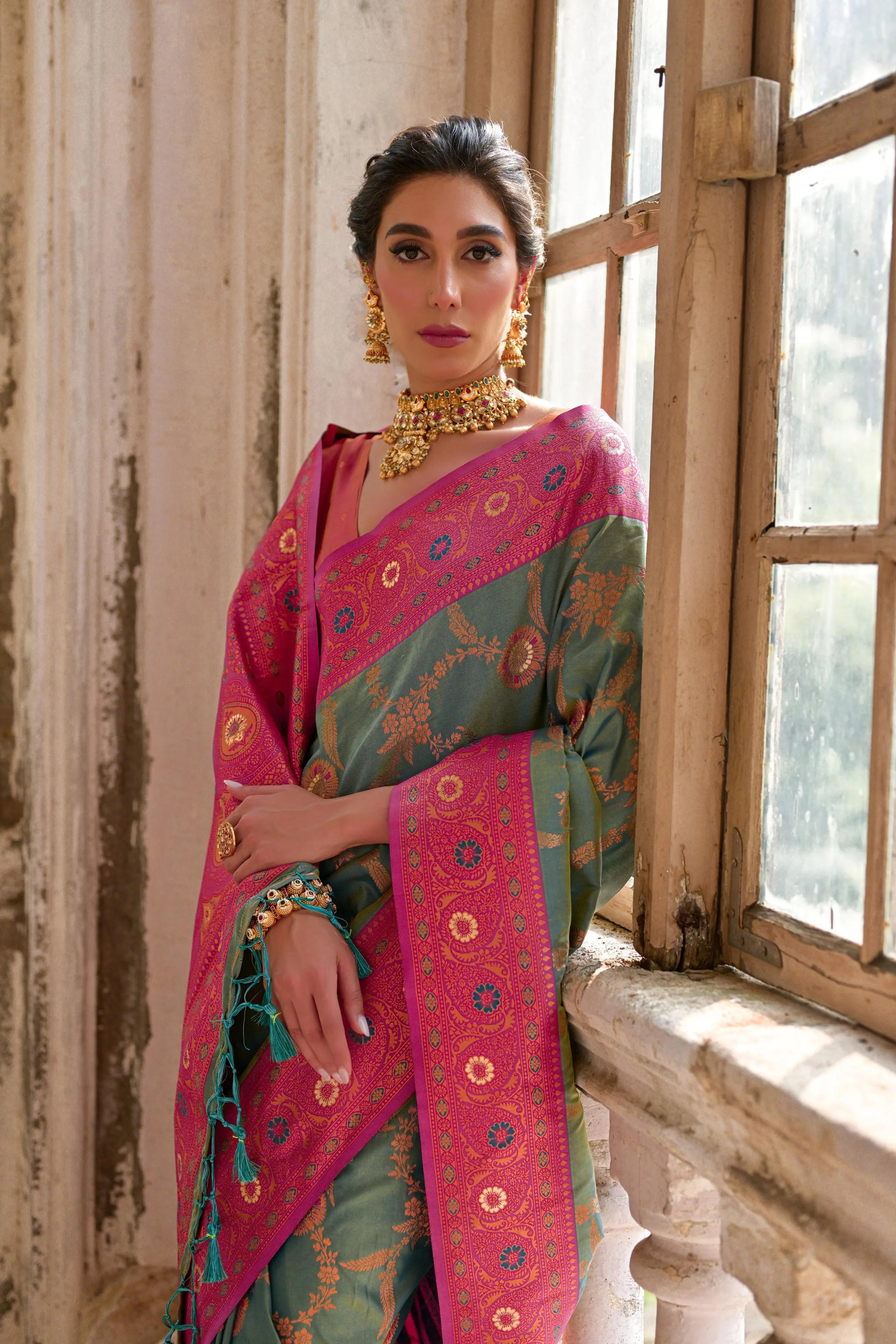 A woman models a striking Dusty Light Green Banarasi Silk Saree with subtle Zari weaving. The saree has a wide, ornate Fuchsia Pink border and pallu featuring dense traditional geometric Zari weaving. She wears a coral pink blouse and heavy gold Kundan jewelry.